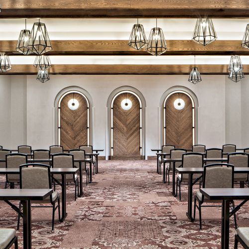 The image shows an empty conference room with several rows of tables and chairs, modern light fixtures, and three decorative wooden doors.