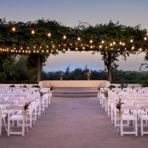 An outdoor wedding venue with white chairs arranged in rows, a floral archway, and string lights overhead at dusk.