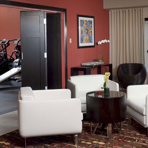 A cozy lounge area with white chairs and a small round table is adjacent to a gym room with exercise equipment in the background.