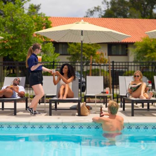 People relaxing by a pool; some on lounge chairs under umbrellas, one in the water, and a server handing drinks to a seated woman.