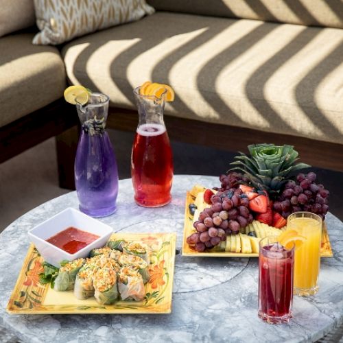 A table with fresh fruit platter, sushi, and various drinks set outdoors on a corner sofa.