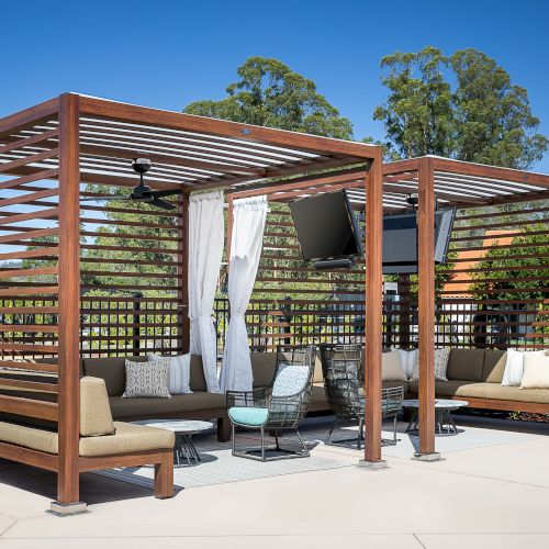 This image shows a modern outdoor seating area with two wooden pergolas, cushioned seating, a TV, and small tables, set on a patio under a clear sky.