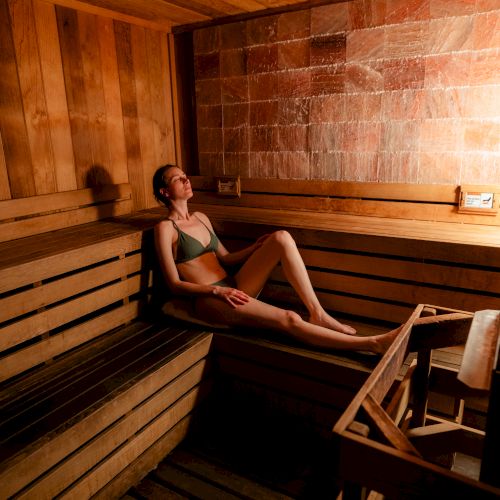 A person relaxing in a wooden sauna, sitting on the bench with knees up, dim warm lighting, and a calm, quiet atmosphere.