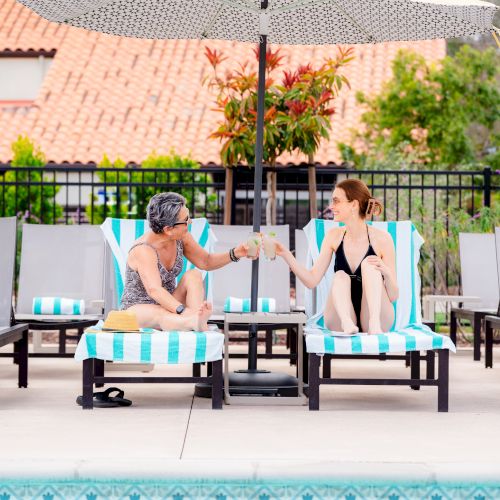 Two people by a poolside, sitting on lounge chairs, sharing a handshake or toast, smiling, with a sunny resort setting behind them.