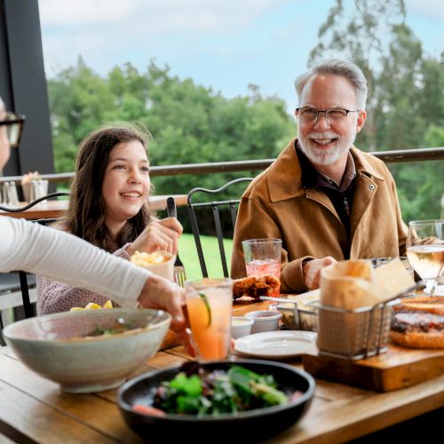 A diverse group enjoys a bright outdoor meal: smiling adults at a wooden table with salads, sandwiches, bowls, drinks, and conversation.
