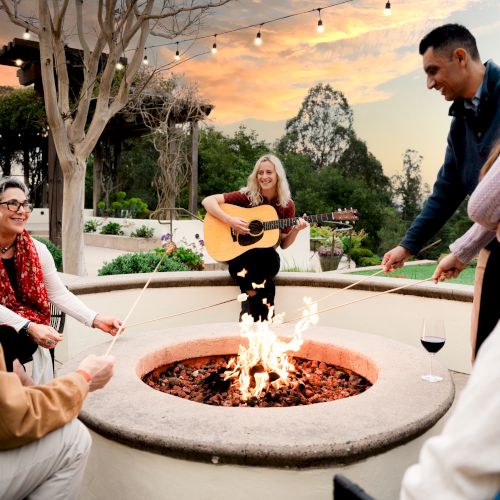 A guitarist warms a small outdoor gathering around a fire pit; friends sit and chat as string lights glow at sunset.