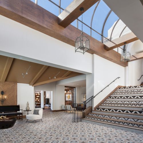 This image shows a spacious, modern lobby with a staircase, skylight, seating area, and patterned tile flooring under natural light from large windows.