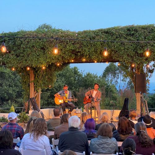 An outdoor concert with two guitarists on stage under a lit pergola, performing for a seated audience, surrounded by lush greenery.