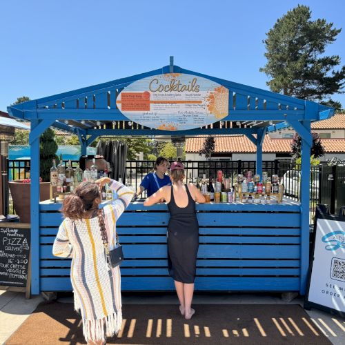 Image shows two people standing by a blue wooden booth labeled "Cocktails"; various bottles are displayed. A signboard and QR code are present.