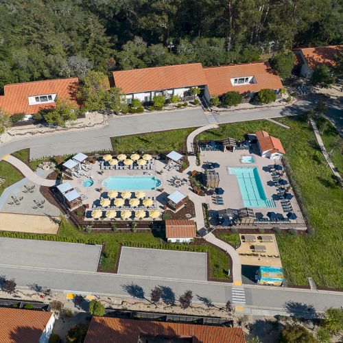 The image shows an aerial view of a resort with multiple buildings, a central pool area, lounge chairs, and surrounding greenery.