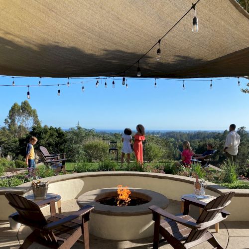 A serene outdoor setting with a firepit, seating, string lights, and people enjoying a scenic view of nature under a sunshade.