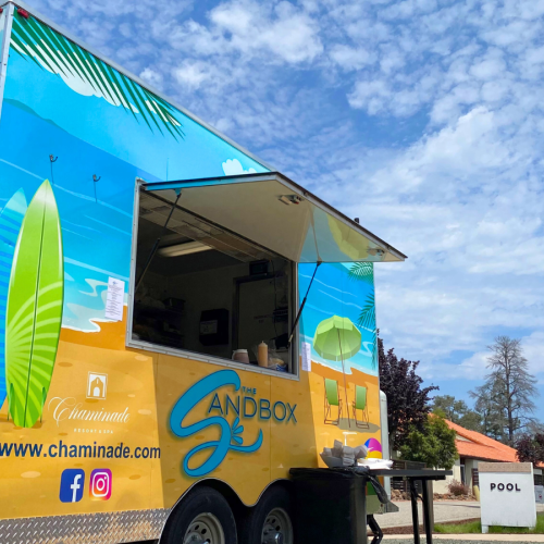 A colorful food truck, labeled "Sandbox," is set up outdoors near a pool, with a bright blue sky and clouds in the background.