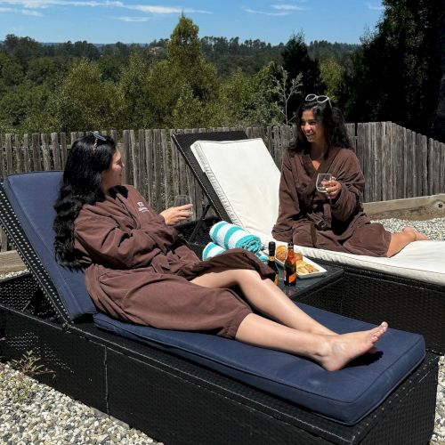 Two people are relaxing on lounge chairs outside, wearing brown robes and talking, with drinks and towels nearby, amidst a scenic, wooded backdrop.