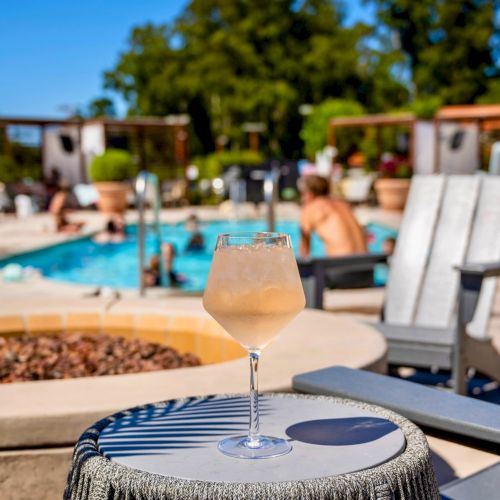 A glass of a light-colored drink on a table with a pool and lounge chairs in the background. People are relaxing by the pool.