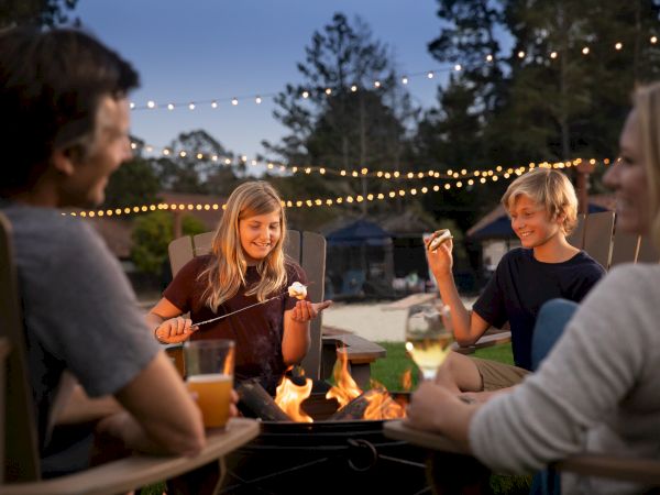 A group of people enjoy roasting marshmallows by a fire pit, surrounded by string lights in an outdoor setting.