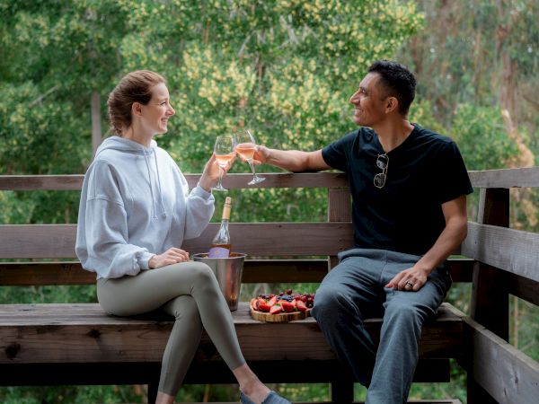 Two people are sitting on a bench, toasting with drinks, outdoors with trees in the background.