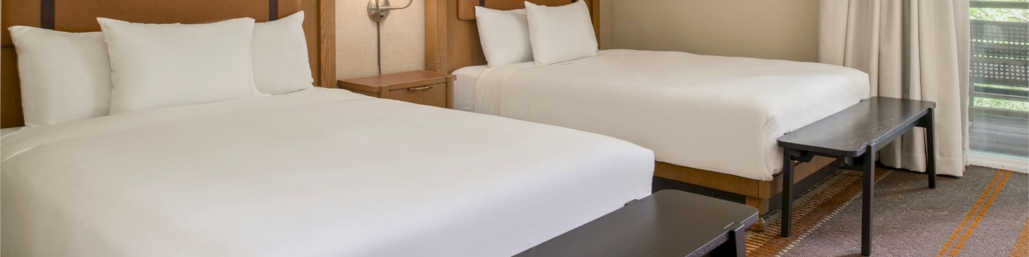 Two queen beds with white linens, wooden headboards, a shared nightstand with a lamp, and a bench at the foot of each bed in a hotel room.