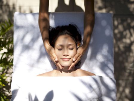 A person is lying down on a white towel outdoors, receiving a facial massage from another person, with shadows of leaves visible.