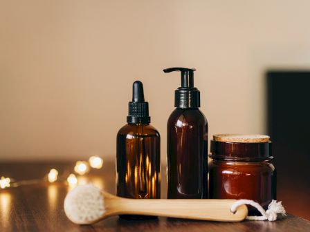 Three brown bottles with dropper, pump, and cork lid, alongside a wooden brush, are placed on a wooden surface with fairy lights in the background.