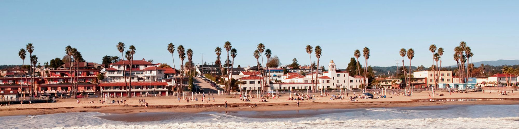 A beach scene with waves crashing, palm trees, buildings in the background, and people walking or relaxing along the shore.