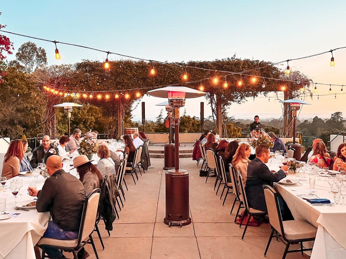A long outdoor rooftop dinner party with string lights, guests seated at white-clothed tables, heaters, and a scenic sunset backdrop.