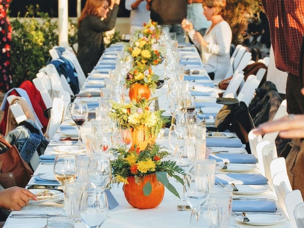 A long table is set for a formal meal outdoors with floral centerpieces in pumpkins, surrounded by people eating and chatting, and wine glasses.