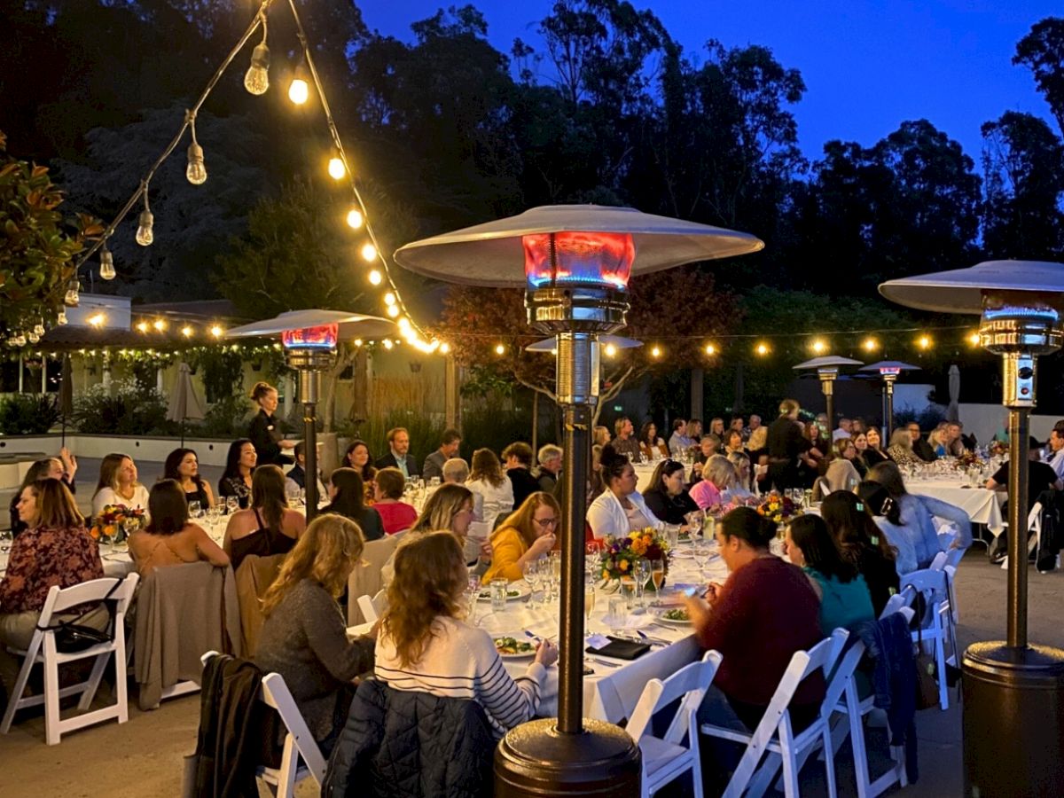 A large group of people are seated around tables at an outdoor evening event, with string lights and patio heaters providing illumination.