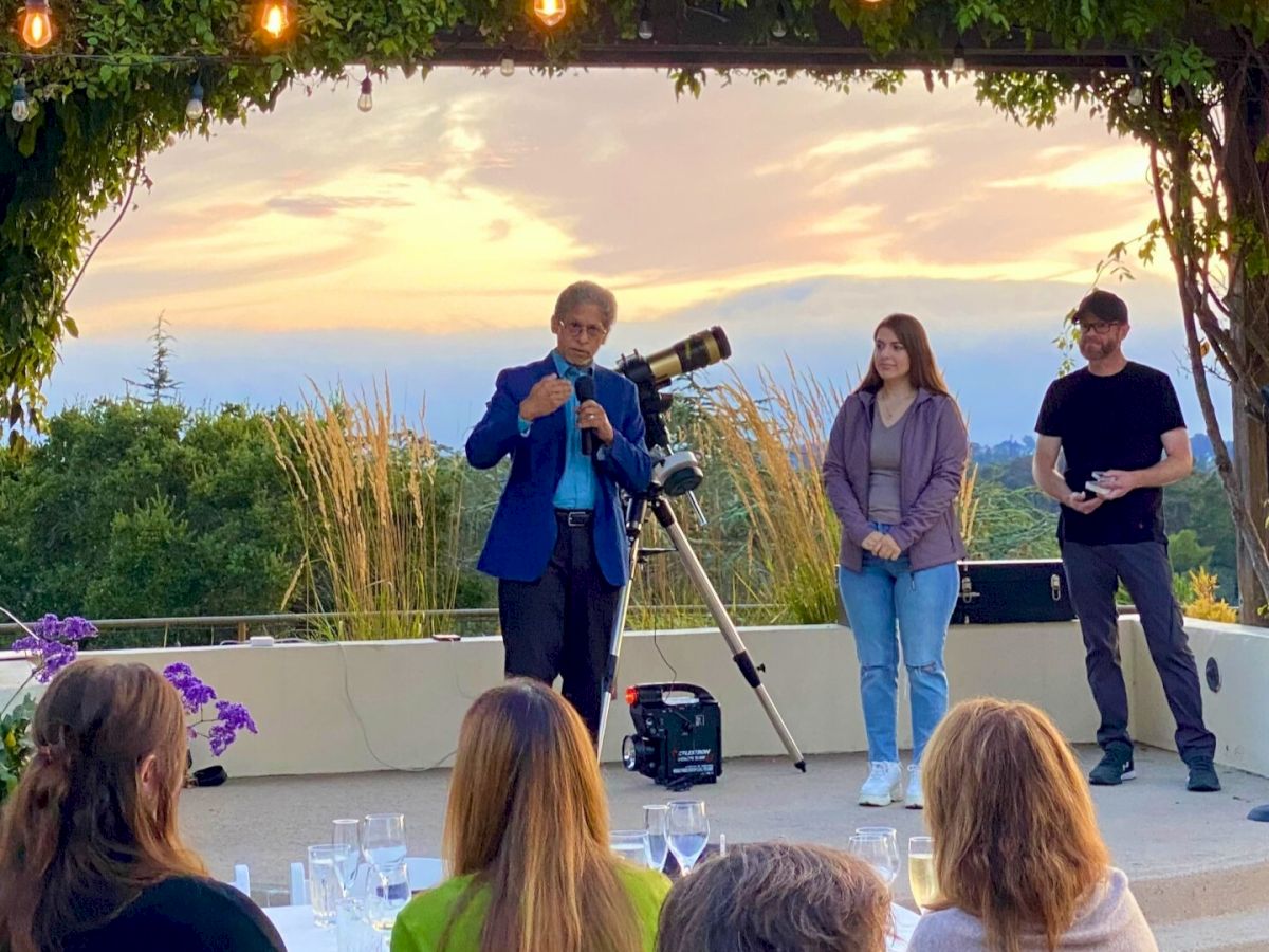 A man speaks to an audience at an outdoor event, with a woman and another man standing nearby and a telescope in the background.