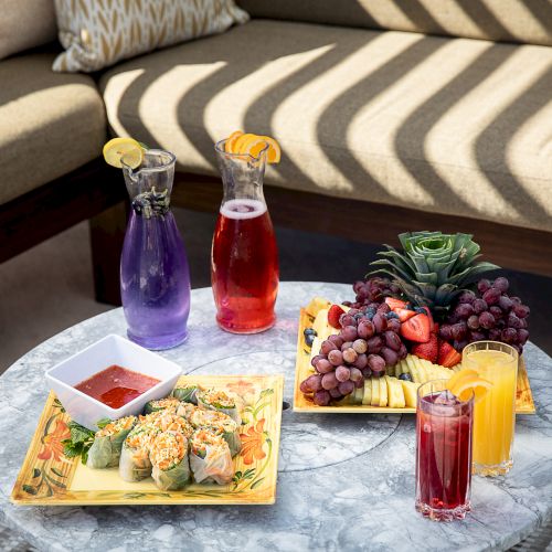 The image shows a table with platters of spring rolls, a variety of fresh fruits, two pitchers of different colored drinks, and three glasses of beverages.