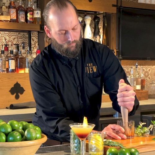 A bartender behind a counter is preparing drinks, with lime and other ingredients around, in a setting decorated with bottles and glasses.