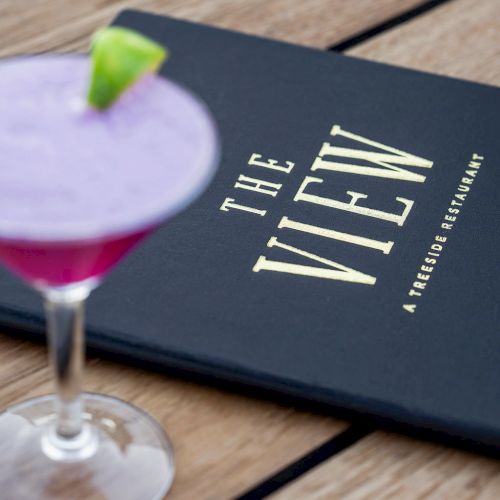 A purple cocktail with a lime wedge and a menu titled "THE VIEW A rooftop restaurant" on a wooden table.