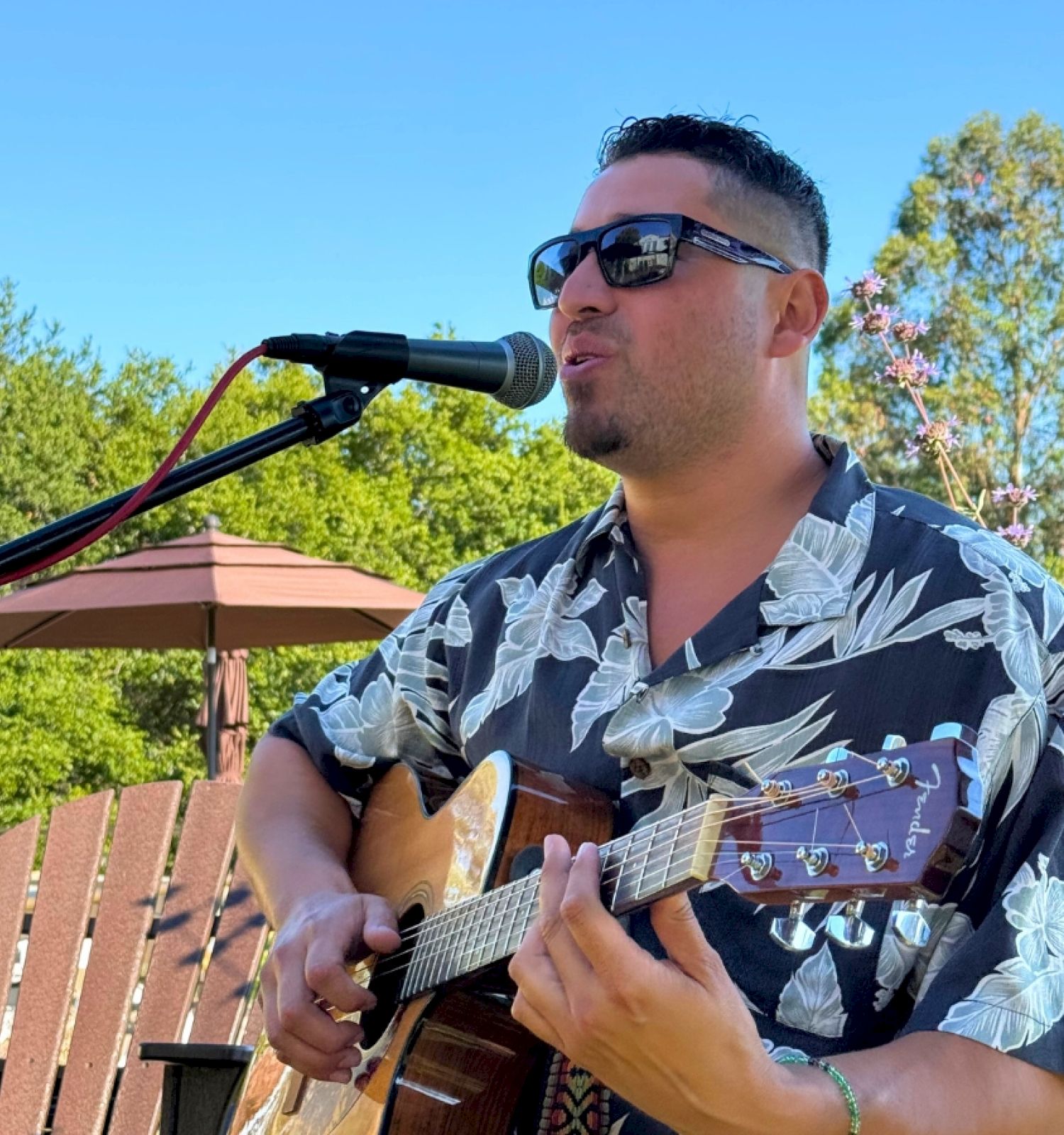 A person wearing sunglasses and a floral shirt plays an acoustic guitar and sings into a microphone outdoors, with trees in the background.