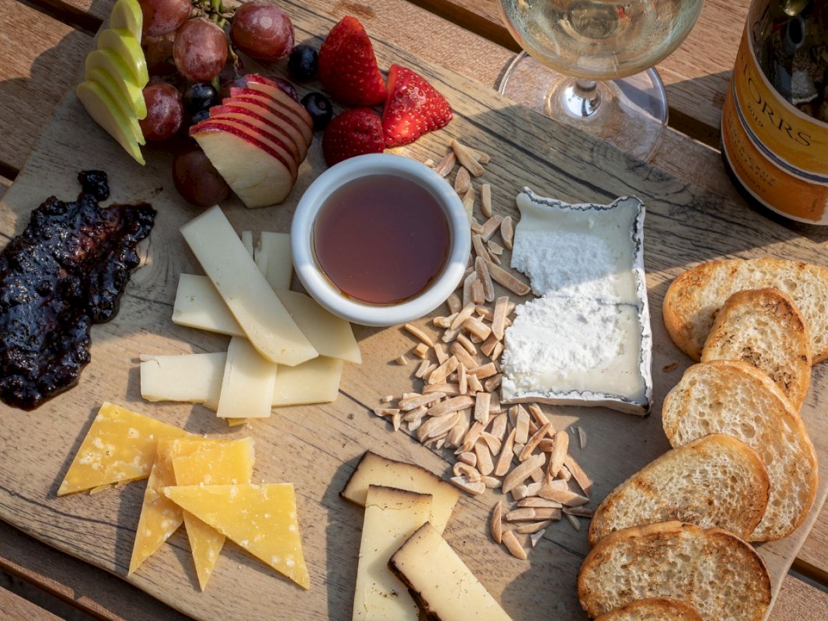 A cheese board with assorted cheeses, fruits, nuts, jam, and bread slices, accompanied by a glass of white wine and a bottle.