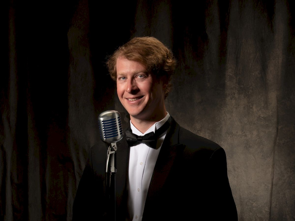 A person in a tuxedo stands in front of a microphone against a dark backdrop, smiling at the camera.