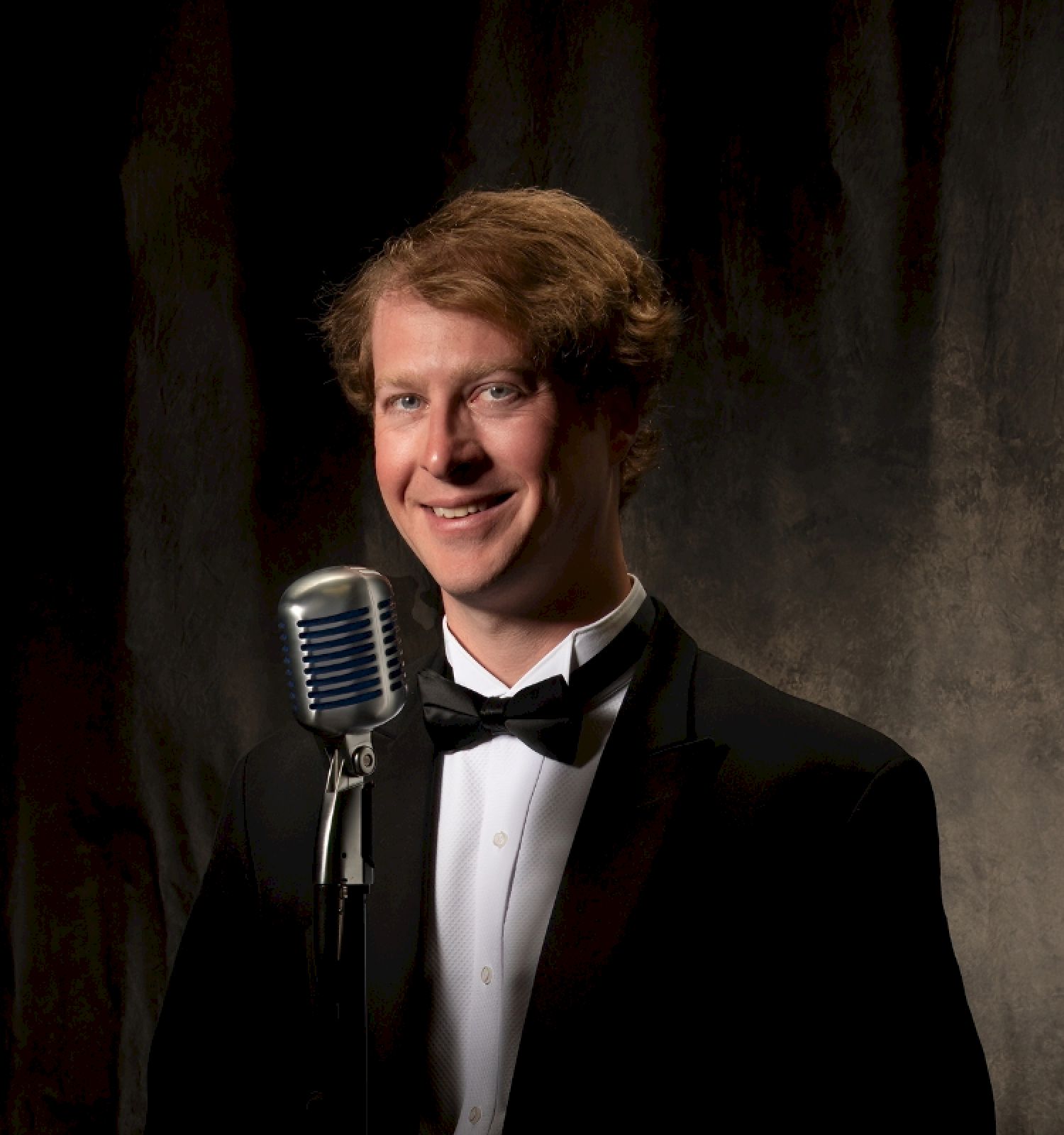 A person in a tuxedo stands in front of a microphone against a dark backdrop, smiling at the camera.