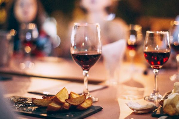 The image shows a table with glasses of red wine, plates of food, and a group of people in a warmly lit setting, possibly a social gathering.