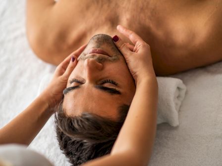 A man is lying down with his eyes closed, receiving a facial massage. His head rests on a towel, and the masseuse's hands cradle his face.