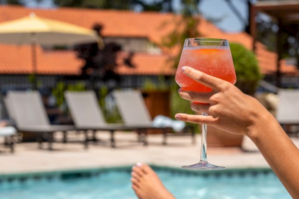 A person relaxes by a pool, holding a glass of a colorful drink, with feet propped up. In the background, lounge chairs and shaded areas can be seen.