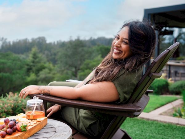 A smiling woman sits in a reclining chair outdoors with a tray of snacks and drinks nearby, relaxing in a scenic garden setting.