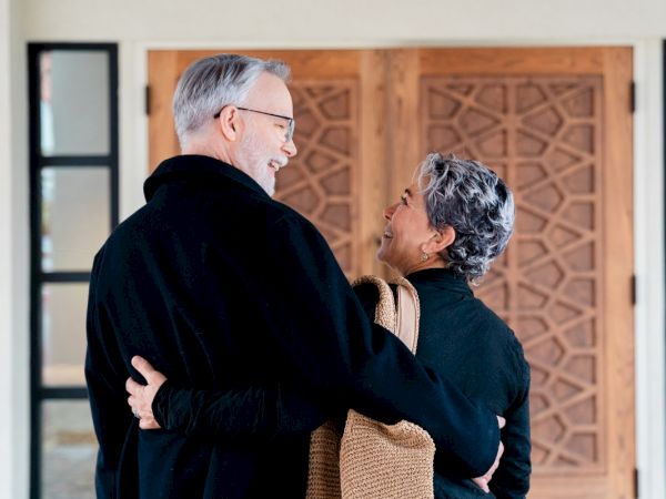 An older couple is gazing at each other and smiling, standing in front of a wooden door, embracing while holding a straw tote.
