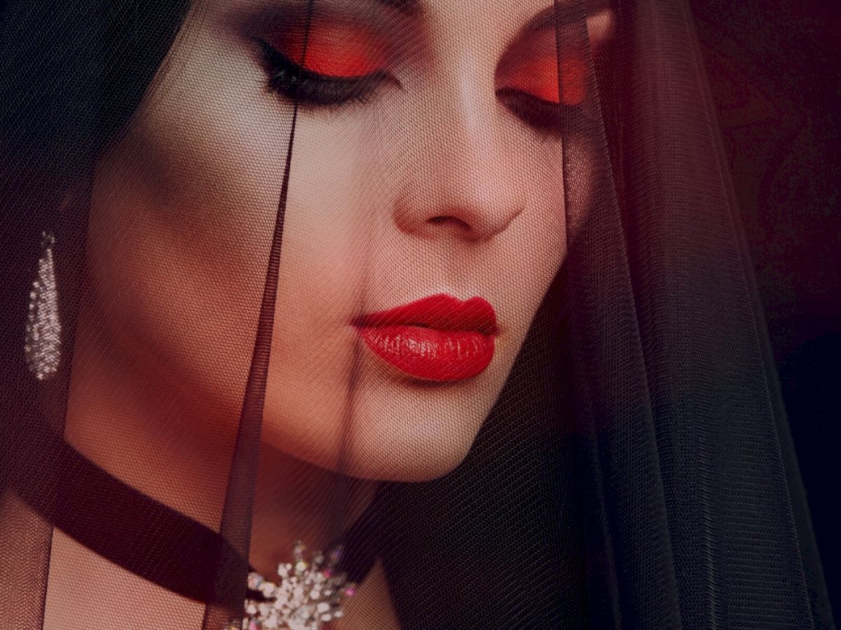 A woman with dramatic red eyeshadow and lipstick, wearing a black veil, diamond earrings, and a jeweled choker.