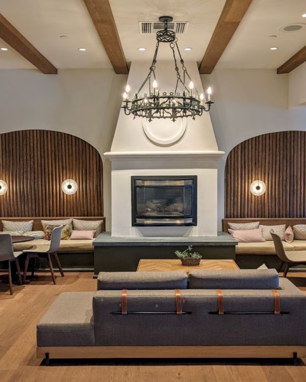 This image shows a modern, cozy lounge with a fireplace, elegant chandelier, wood accents, and comfortable seating areas.