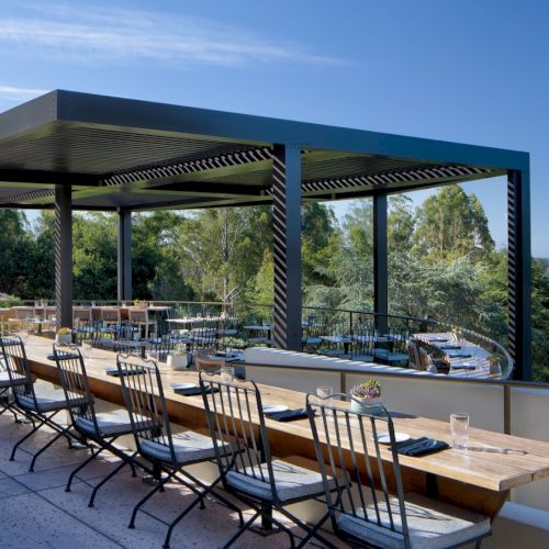 This image shows an open-air dining area with long wooden tables and chairs, partially covered by a pergola, with a view of trees and nature beyond.