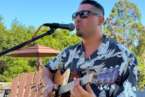 A man wearing sunglasses and a floral shirt is playing an acoustic guitar and singing into a microphone outdoors under a clear blue sky.