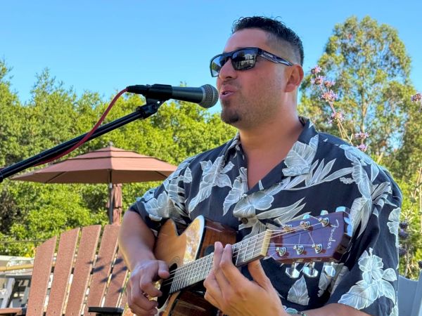 A man wearing sunglasses and a floral shirt is playing an acoustic guitar and singing into a microphone outdoors under a clear blue sky.
