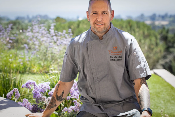 A chef wearing a grey uniform stands outside with a scenic background of greenery and purple flowers, smiling at the camera.