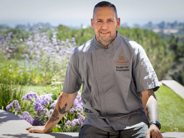 A chef wearing a grey uniform stands outside with a scenic background of greenery and purple flowers, smiling at the camera.