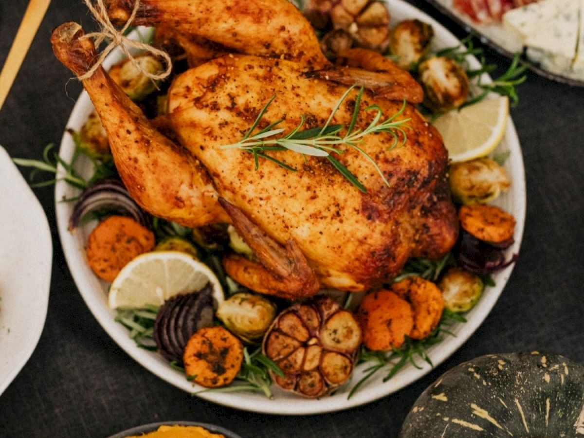 A roasted chicken garnished with herbs and surrounded by vegetables, accompanied by a plate of sliced meats and cheeses, and a bowl of orange sauce.