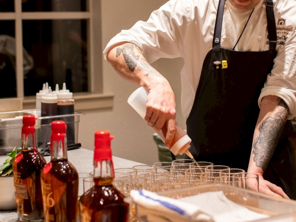 A person in a chef's uniform is pouring liquid into glasses. There are bottles with red caps on the counter.