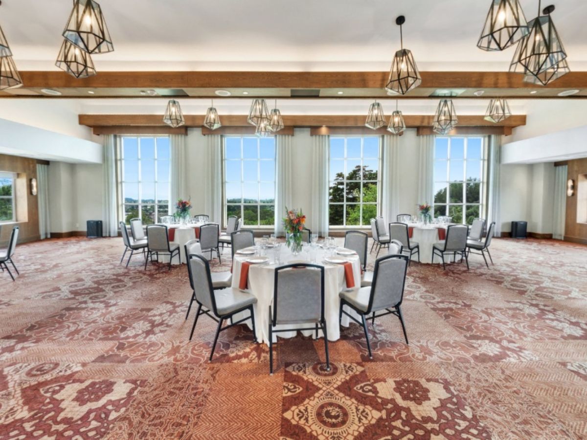 A spacious, elegant banquet room with round tables, chandeliers, large windows, and a patterned carpet, set for an event or gathering.